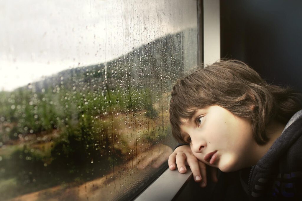 Junge schaut gelantweilt aus dem Fenster in die Landschaft bei Regen.